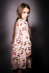 A young girl with long brown hair is looking over her right shoulder at the camera. She is wearing a light pink dress with colorful printed designs of animals and scooters against a dark, neutral background. The lighting highlights her face and dress patterns, capturing a beautiful moment of family photography.