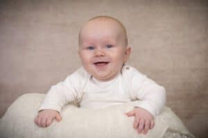 A happy baby with blue eyes and a light complexion is smiling while lying on a soft white blanket. The baby is wearing a long-sleeved, light-colored onesie. The background is a warm beige color.