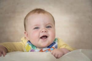 A baby with light brown hair wearing a colorful bib and a yellow shirt smiles joyfully while leaning on a soft surface. The background is a neutral tone.