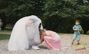A bride in a white gown and veil bends over as a woman in a pink dress adjusts something near her feet. A young boy in a green shirt and shorts stands nearby, watching. They are outdoors on a path surrounded by greenery.