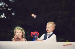 A young boy in a vest and bow tie uses a toy to launch an object into the air, while a girl with a flower crown and a light pink dress blows a kiss. They are seated at a white table with a dark background. Text on the image reads, "Tim Sutton Photography.