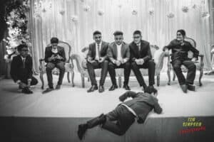 A group of young men dressed in formal attire sit on a decorated stage. One boy is kneeling and looking at his phone while another boy lies on his stomach near the edge of the stage. Flower arrangements are visible in the background. The image is in black and white.