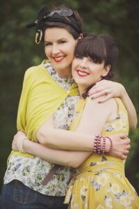 Two women, smiling and hugging each other, are dressed in vibrant vintage-style clothing. One wears a yellow shawl and the other a yellow flower-patterned dress. Both have dark hair, and their bright red lipstick complements their cheerful expressions. The green, leafy background adds a charming touch to this heartwarming moment of vintage photoshoot in Saddleworth