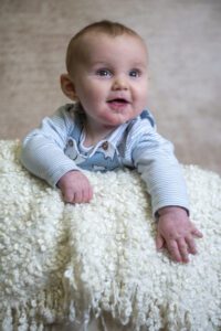 A baby wearing a striped onesie and overalls leans on a soft, textured, cream-colored blanket. The baby is looking slightly upward with a content expression, mouth slightly open, with a small drool spot visible on their chin.