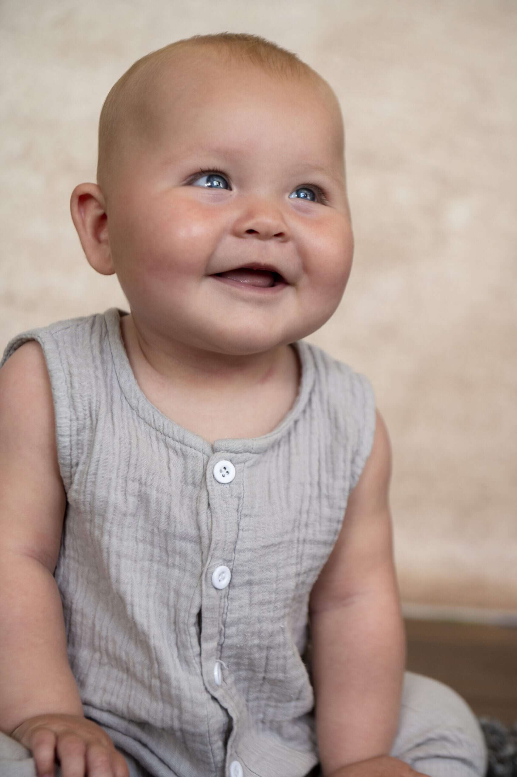 A baby with light skin and blonde hair is sitting and smiling. They are wearing a sleeveless, light gray, buttoned outfit and looking slightly upwards. The background is a soft beige color.