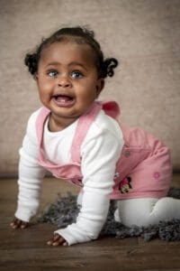 A baby with curly hair crawls on a wooden floor. The baby is dressed in a white long-sleeve shirt, a pink overall dress with a character printed on it, and white tights. The baby has an expressive face and is looking slightly upward. The background is neutral-colored.