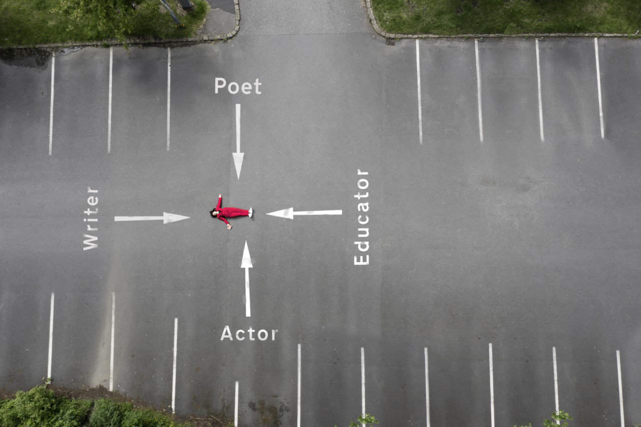 lisa moore in red lies on their back in the middle of an empty parking lot, framed by the greenery of Saddleworth. Four large white arrows on the ground point in different directions, labeled "Writer," "Poet," "Educator," and "Actor." It's a scene that could be straight out of brand photography.