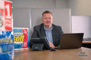 A man in a gray suit jacket sits at a desk with a laptop and phone in front of him. He is smiling and has short hair. On the left side of the desk, there are packages of toilet paper—part of a brand photography shoot. The background is a neutral office setting with a TSP Studios logo in the lower right corner.