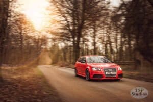 A red Audi RS3 speeds down a country road flanked by barren trees. The sun sets in the background, casting a warm glow over the scene. A TSP Studios watermark is in the bottom right corner.