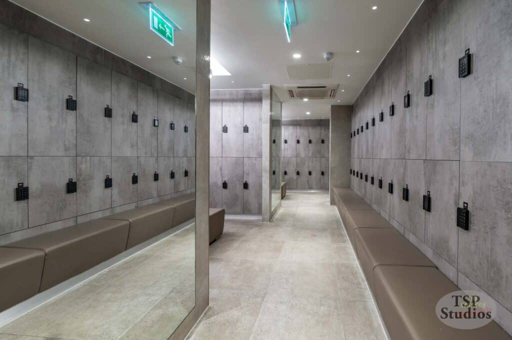 A modern locker room with gray concrete walls and lockers featuring digital keypads. Tan upholstered benches run along both sides of the room. The ceiling has recessed lighting and an emergency exit sign glows in the background. "TSP Studios" logo is visible in the corner.