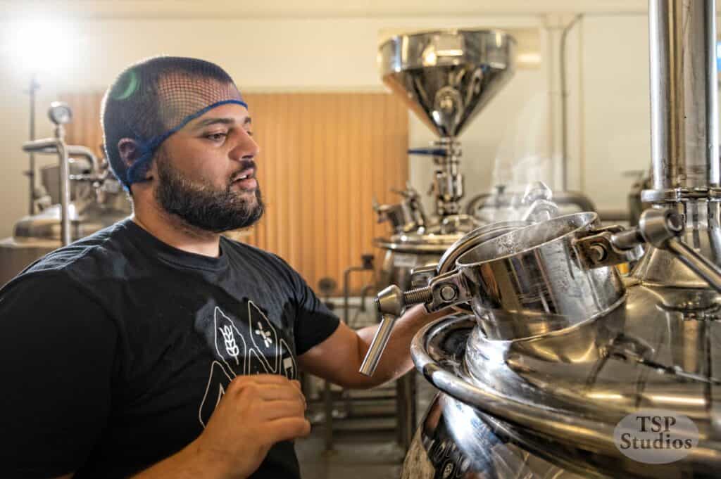 A bearded man wearing a hairnet on his head and a black shirt with a wheat graphic is working in a brewery. He is looking attentively at a metal vat with steam rising from it, surrounded by brewing equipment. The logo "TSP Studios" is visible in the bottom right.