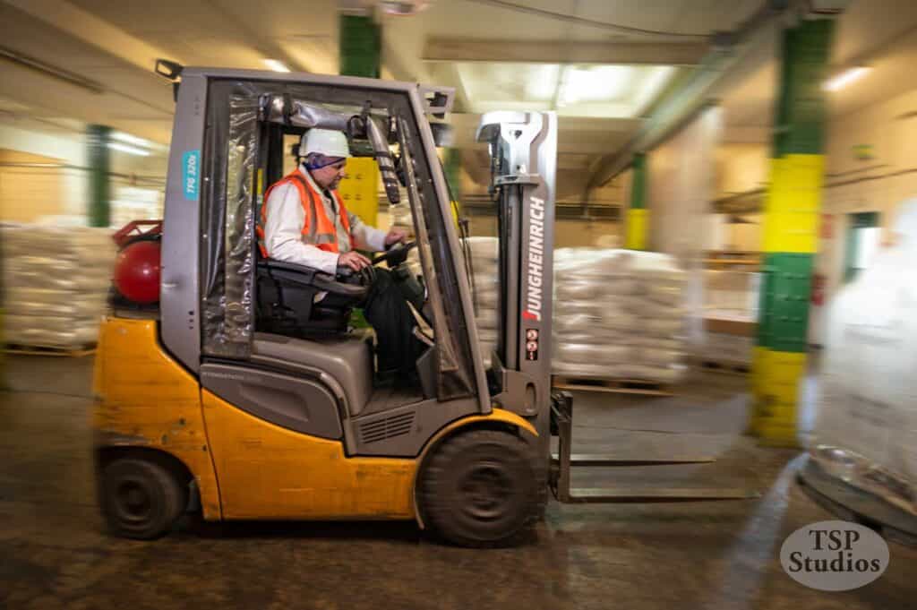 A worker wearing a white helmet, orange safety vest, and protective glasses operates a Jungheinrich forklift in an industrial warehouse. They are transporting goods wrapped in clear plastic. The environment features large bags and pillars painted in green and yellow.