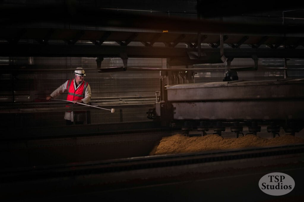 A worker wearing a white helmet and safety vest operates machinery in an industrial setting. They hold a long tool and are surrounded by dark, industrial equipment with piles of processed material. The logo "TSP Studios" appears in the bottom-right corner.
