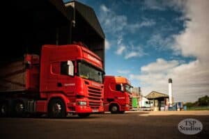 Two red Openfield trucks are parked in front of a large industrial warehouse under a partly cloudy sky. The scene includes additional industrial buildings and equipment in the background. The logo "TSP Studios" is visible in the bottom right corner.