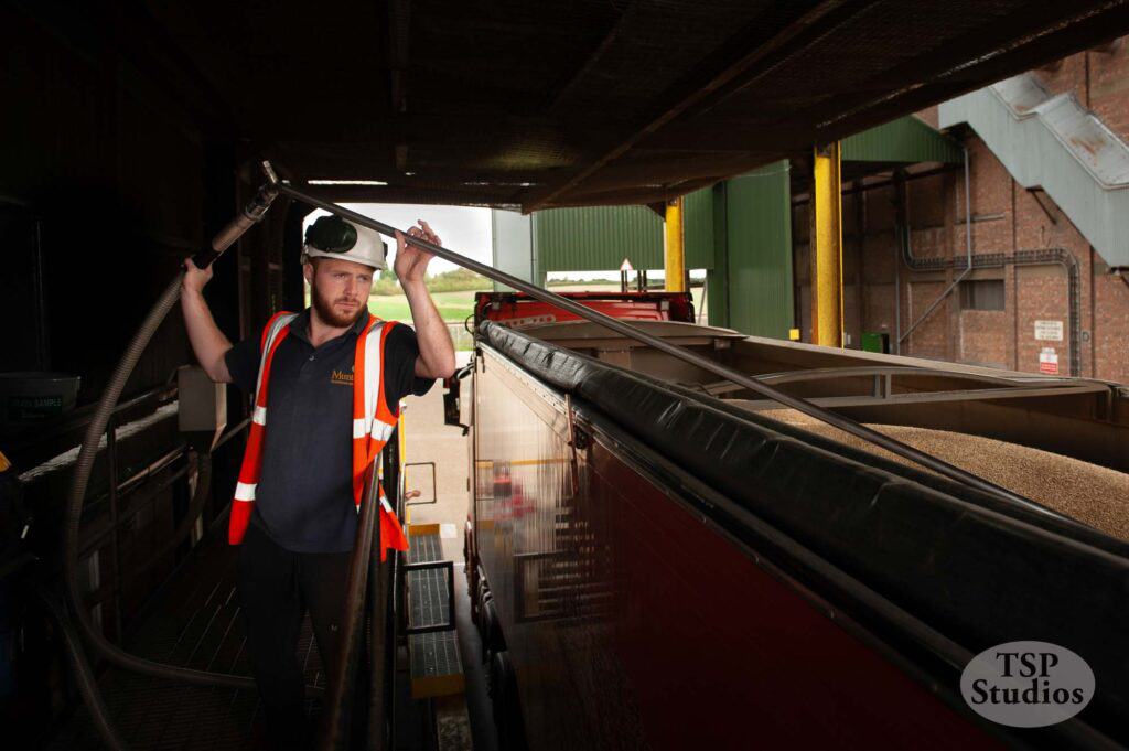 A worker in a high-visibility vest and hard hat operates a large hose, loading grain into a long red truck parked at an agricultural facility. The scene appears to be an indoor area with parts of machinery and structures in the background. The image is marked "TSP Studios.