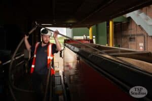 A worker in a high-visibility vest and hard hat operates a large hose, loading grain into a long red truck parked at an agricultural facility. The scene appears to be an indoor area with parts of machinery and structures in the background. The image is marked "TSP Studios.