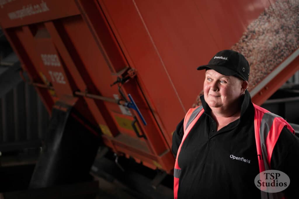 A person wearing a black cap and a black shirt with the word "Openfield" stands in front of a large red industrial machine, possibly a grain trailer. The person is also wearing a high-visibility pink safety vest. The photo is labeled with a "TSP Studios" logo.