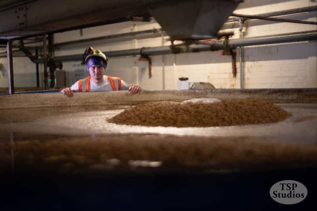 A person wearing a hard hat and safety vest inspects a large tank filled with bubbling liquid and a pile of solid material at an industrial facility. Pipes and equipment are visible in the background. The image is branded with a "TSP Studios" logo.