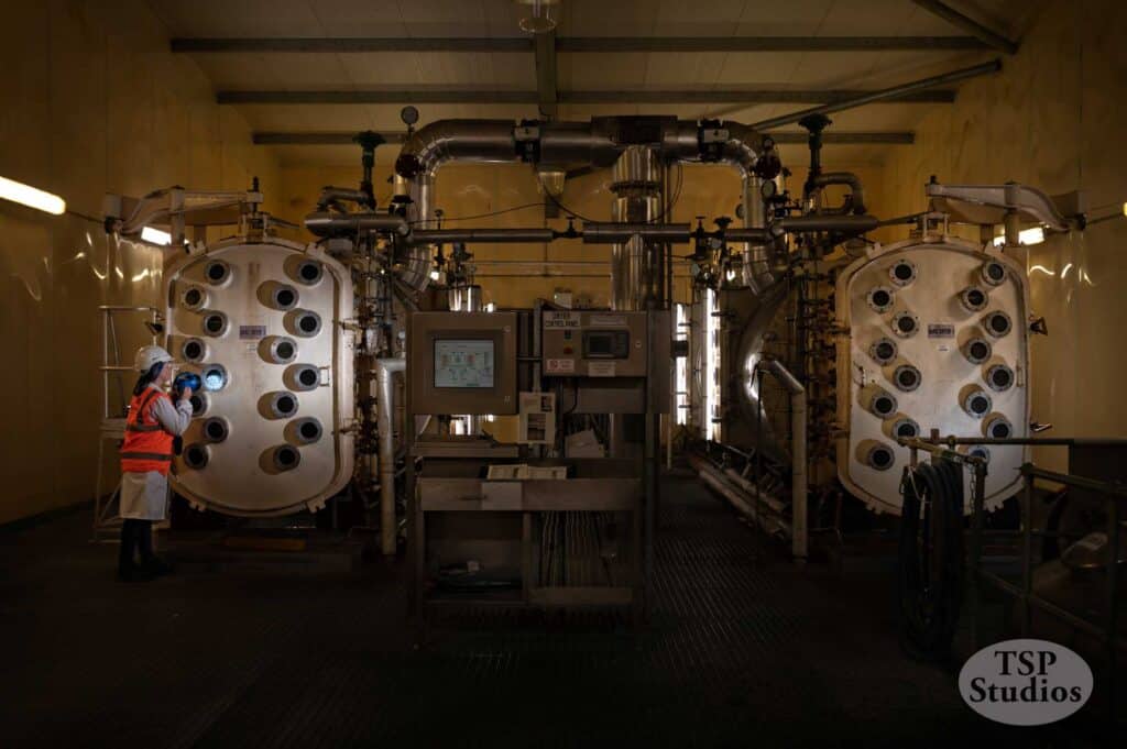 A worker in high-visibility gear and a hard hat examines one of two large industrial machines in a dimly lit facility. The machines both have multiple round openings. A control panel sits between them. TSP Studios logo is present at the bottom right.