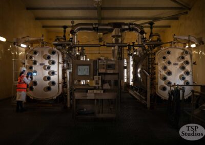A worker in high-visibility gear and a hard hat examines one of two large industrial machines in a dimly lit facility. The machines both have multiple round openings. A control panel sits between them. TSP Studios logo is present at the bottom right.