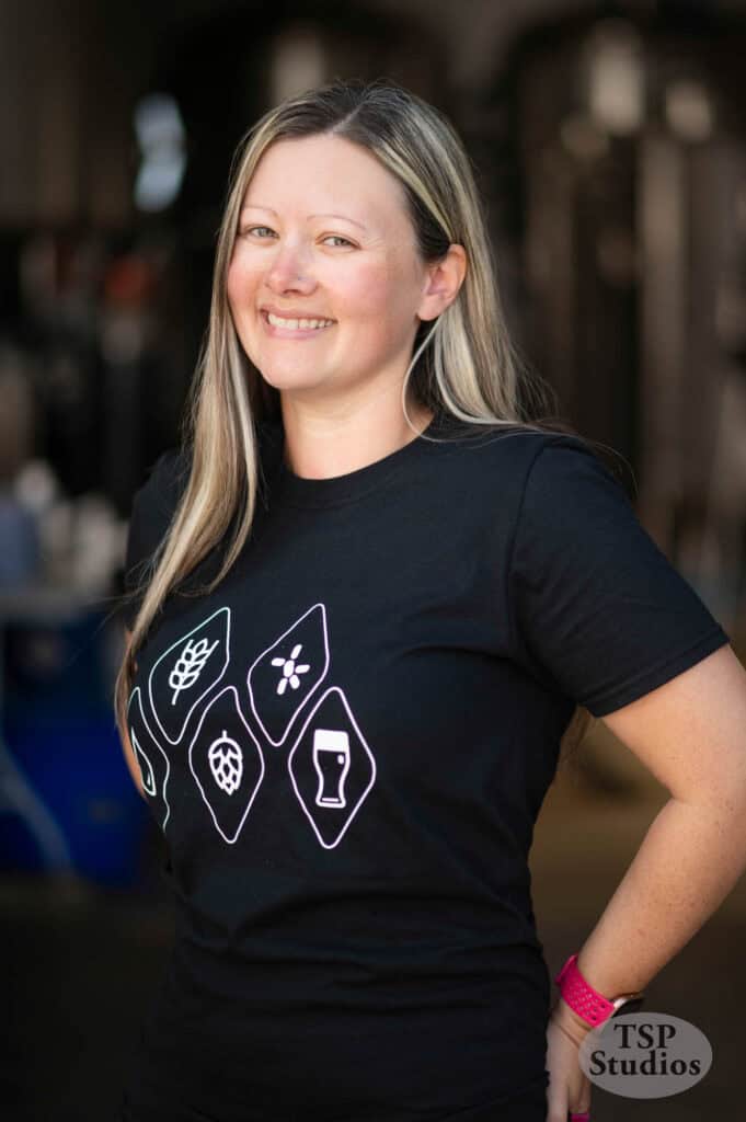 A person with long, blonde hair is smiling and standing in a dimly lit indoor setting. They are wearing a black T-shirt with white illustrations of hops, a flower, a plant, and a beer glass. The logo "TSP Studios" is visible in the bottom right corner.