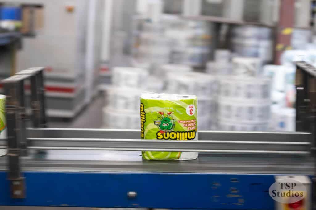 A brightly colored green and yellow package of toilet paper with cartoon graphics moves along an industrial conveyor belt in a manufacturing plant. The background is blurred, showing various equipment and more packaged products. The TSP Studios logo is in the bottom right.