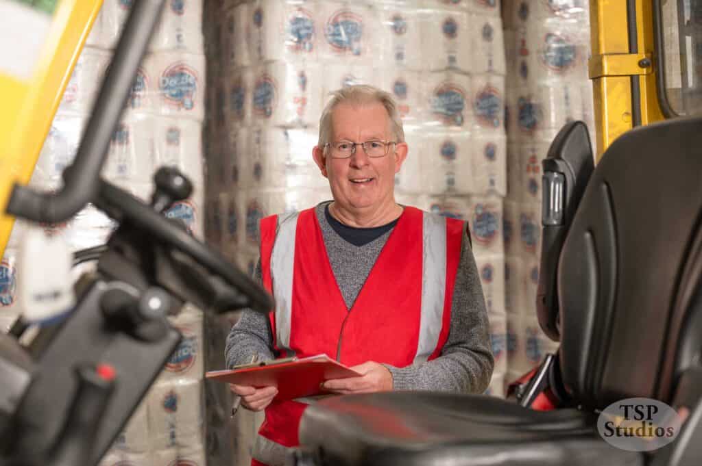 An elderly man wearing a red safety vest and glasses is holding a clipboard and smiling. He is standing in what appears to be a warehouse filled with stacked packages of toilet paper. The image includes a piece of machinery in the foreground.