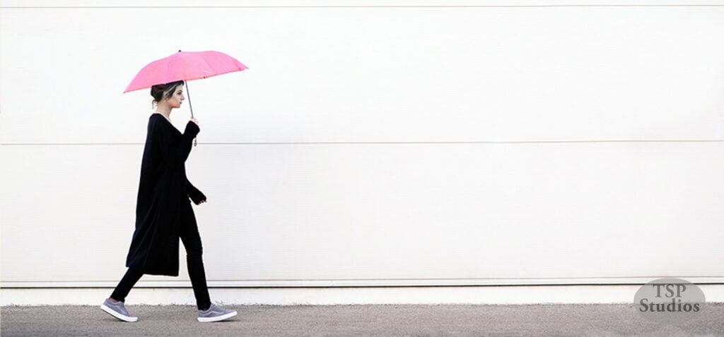 A person dressed in black walks on a sidewalk while holding a pink umbrella. The background features a plain, light-colored wall with the TSP Studios logo in the bottom right corner.