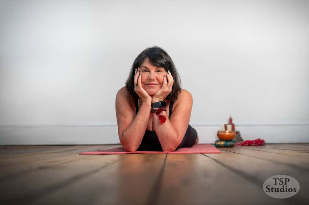 A woman lays on a yoga mat, resting her head on her hands with a serene expression. She's in a room with wooden floors and a plain white wall. Nearby, there are some yoga props, including a small statue and a few colorful items. The logo "TSP Studios" is in the corner.