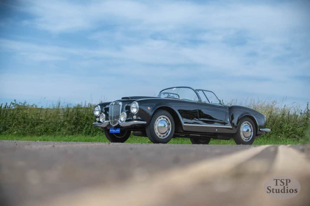 A classic black convertible car with chrome accents is parked on a country road under a blue sky with scattered clouds. The car’s sleek design is highlighted by the low-angle shot. Tall grass lines the road, adding a natural backdrop. The photo includes a watermark reading "TSP Studios" at the bottom-right corner.