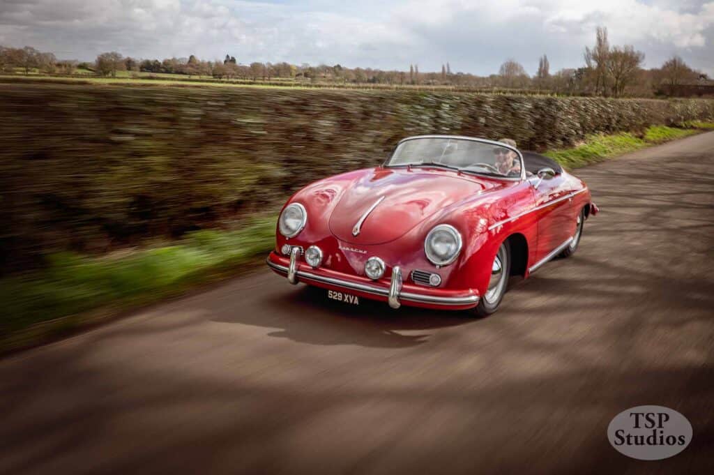A classic red convertible sports car with a license plate reading "529 XYA" is speeding down a rural road. The background features blurred greenery and trees, indicating motion. The image is credited to TSP Studios in the bottom right corner.