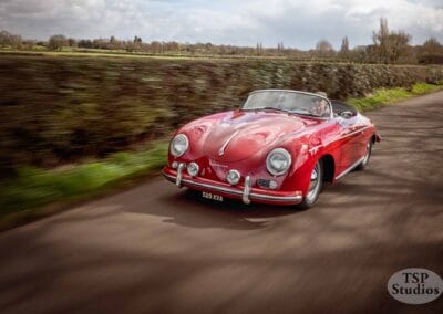 A classic red convertible sports car with a license plate reading "529 XYA" is speeding down a rural road. The background features blurred greenery and trees, indicating motion. The image is credited to TSP Studios in the bottom right corner.