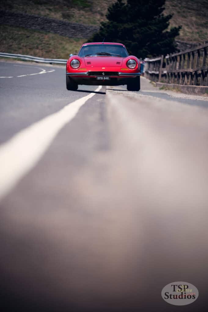 A red vintage sports car is parked on the side of an empty, curving road. The perspective is from ground level, focusing on the car in the distance with the road's white center line leading up to it. There are wooden guardrails on the right side.