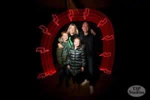 A family of four, consisting of two adults and two children, stand together smiling in the dark. They wear winter jackets, and an arch of red light streaks forms a frame around them. The logo “TSP Studios” is displayed in the lower right corner.