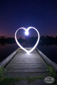 A glowing heart shape, reminiscent of creative mobile photography, appears above a wooden dock at night, with a calm lake and tree silhouettes in the background. The sky is dusky purple. "TSP Studios" is written in the bottom right corner.
