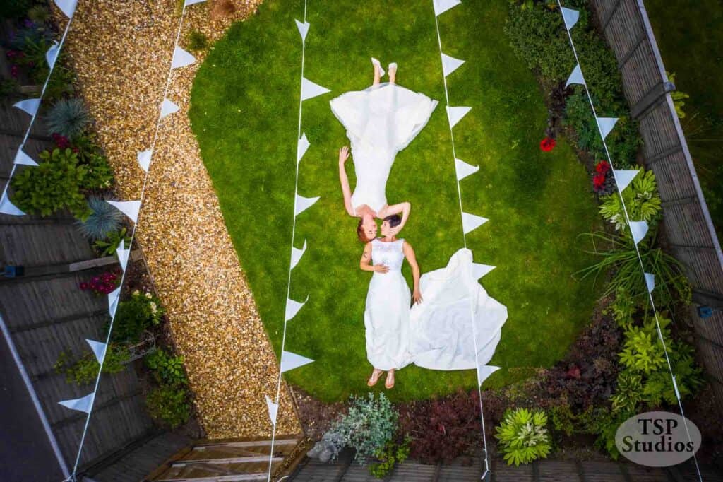 Two women in wedding dresses lie side by side on a patch of green grass, nestled in a garden. Festoons with white triangular flags hang above them, framing the scene. The women hold hands, and the surrounding area features gravel paths and various plants. TSP Studios watermark is in the bottom-right corner.