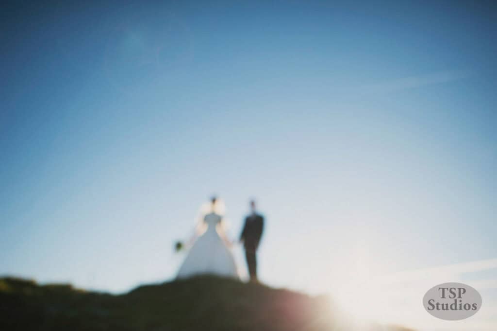 Silhouetted against a bright blue sky with lens flare, a blurry image shows a bride in a gown and groom in a suit holding hands on a hill. The sun is low, casting light from the horizon. "TSP Studios" is visible in the bottom right corner.
