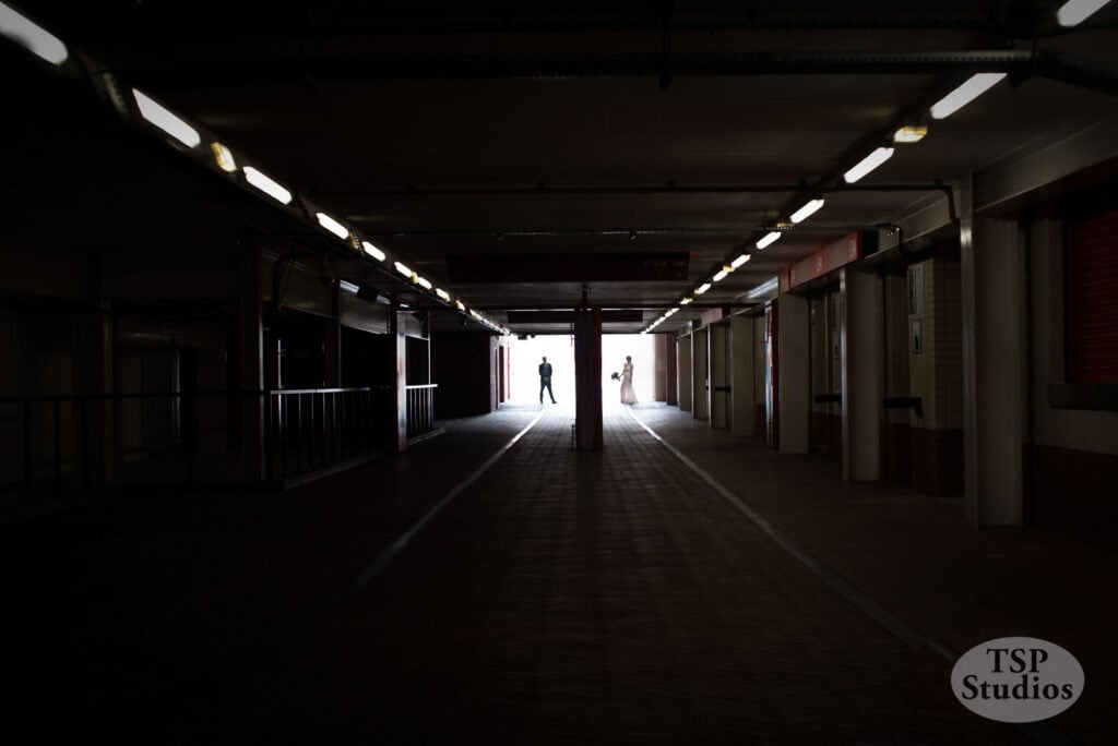 A dimly lit tunnel with illuminated fluorescent lights on the ceiling and an open, bright exit at the end. Two distant figures are walking towards the exit, silhouetted against the light. The TSP Studios logo is in the bottom right corner.