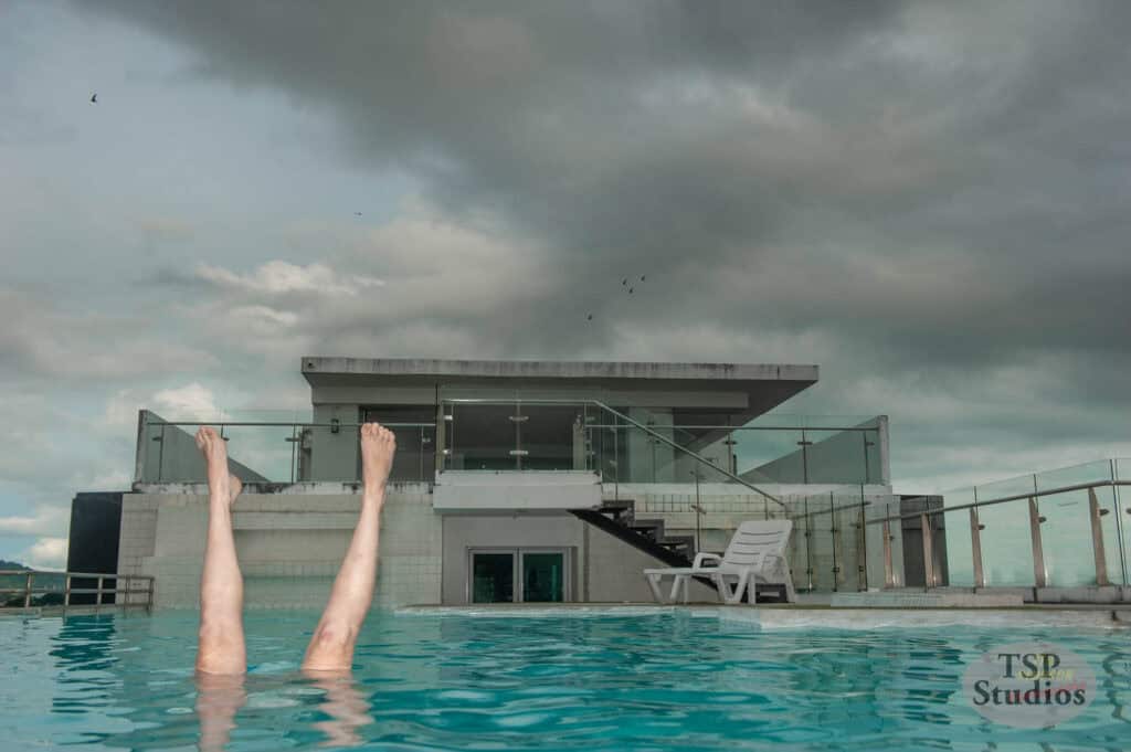 A person is floating on their back in a swimming pool, with only their legs sticking straight up out of the water. The pool is on a rooftop, with a modern building in the background and a cloudy sky overhead. An empty lounge chair and a staircase are also visible.
