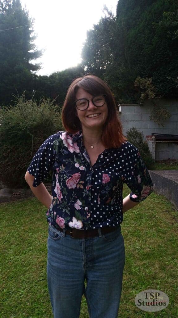 A person with shoulder-length brown hair and glasses is standing outdoors on grass. They are smiling and wearing a floral and polka dot shirt with blue jeans. Trees and bushes are visible in the background. The bottom right corner has a watermark that reads "TSP Studios.