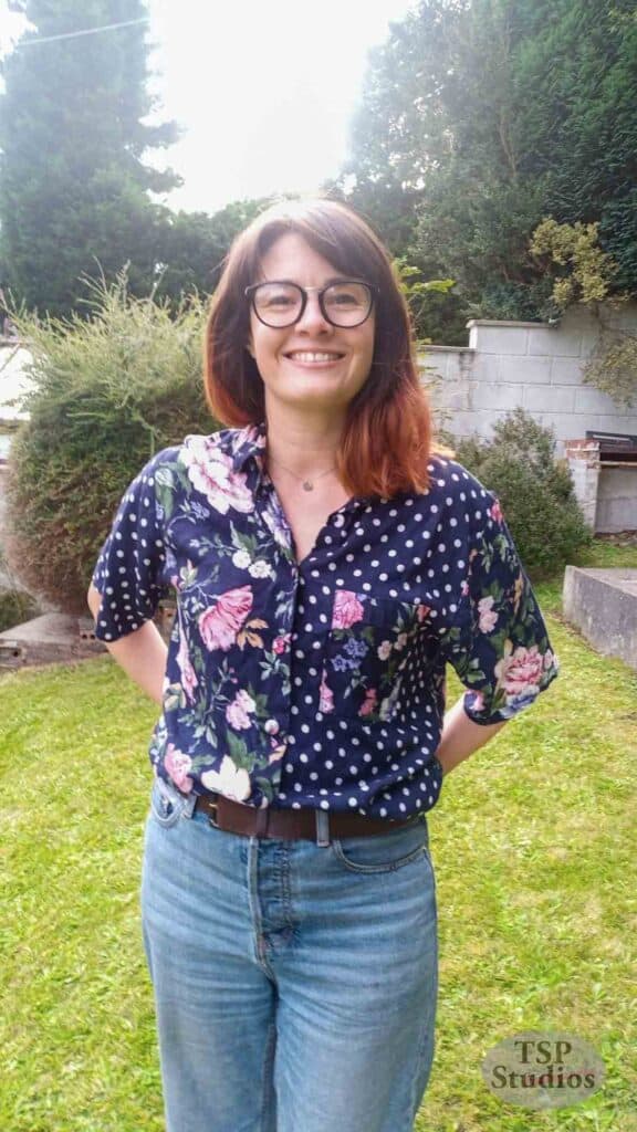 A person with shoulder-length hair and glasses stands outdoors on grass, smiling at the camera. They wear a blue shirt with a mixed floral and polka dot pattern, tucked into high-waisted jeans. Shrubs and trees are visible in the background. Text at the bottom reads "TSP Studios.