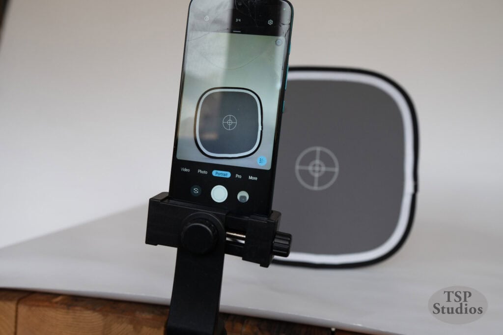 A smartphone on a tripod stand is capturing an image of a gray and white focus target placed in the background. The screen of the phone shows the target in focus. The logo "TSP Studios" is visible in the bottom right corner of the image.