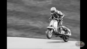 A black and white photo of a motorcyclist performing a jump on a vintage scooter. The rider, dressed in racing gear and helmet, is airborne with the scooter tilted slightly forward, against a blurred background. The logo "TSP Studios" is visible in the bottom right corner.