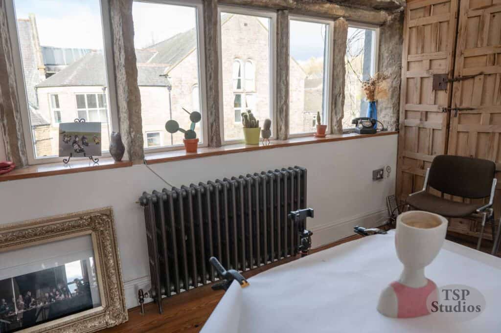 A room with large multi-paned windows features an assortment of potted plants on the windowsill. There's a framed picture leaning against the wall, a radiator under the windows, and a chair in the corner. In the foreground, a table holds an unfinished sculpture.