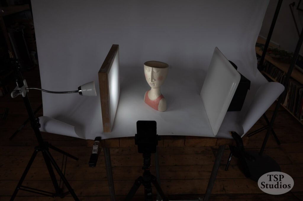 A mannequin head is positioned in the center of a studio setup. It is illuminated by a desk lamp on the left and flanked by reflective surfaces on either side to control lighting. The scene is arranged on a wooden table against a white background.