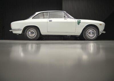 Side view of a vintage white alfa romeo giuila car with chrome detailing and a green emblem on the front fender, parked indoors against a dark, textured wall. The polished floor reflects the vehicle's streamlined design.