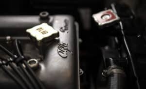 Close-up of a vintage engine showing an "olio" cap and the engraved name "Alfa Romeo." Visible wires and engine components add to the industrial feel. The lighting highlights the metal texture.