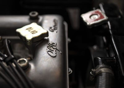 Close-up of a vintage engine showing an "olio" cap and the engraved name "Alfa Romeo." Visible wires and engine components add to the industrial feel. The lighting highlights the metal texture.