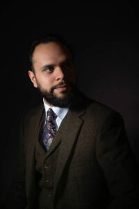 A man with a beard is wearing a dark green suit, white shirt, and patterned tie. He is gazing thoughtfully to the side against a dark background, with soft lighting highlighting his face.