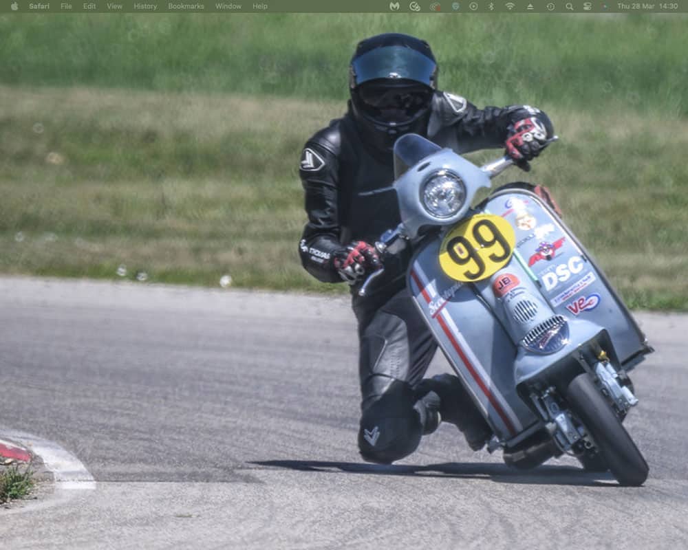 Tim Simpson Scooter Racing A lambretta leans into a turn on a race track. The motorcycle has the number 99 on the front. The background shows a grassy field blurred by motion.